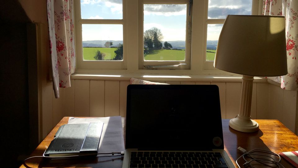 A laptop and notebook on a desk facing a view over green fields and trees on a sunny day