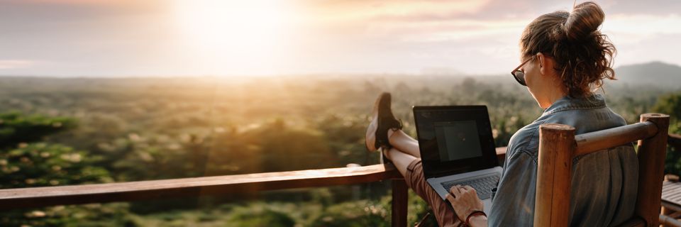 A woman works on her laptop sitting on a balcony, with a beautiful view of a sunrise over the countryside
